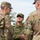 Three military personnel are engaged in a conversation outdoors. Two individuals wear U.S. Army OCP (Operational Camouflage Pattern) uniforms with American flag patches, while the central figure wears a Japan Ground Self-Defense Force (JGSDF) uniform with a name tag reading 'K. TANAKA'. This appears to be a joint military exercise or liaison meeting between U.S. and Japanese armed forces.