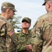 Three military personnel are engaged in a conversation outdoors. Two individuals wear U.S. Army OCP (Operational Camouflage Pattern) uniforms with American flag patches, while the central figure wears a Japan Ground Self-Defense Force (JGSDF) uniform with a name tag reading 'K. TANAKA'. This appears to be a joint military exercise or liaison meeting between U.S. and Japanese armed forces.
