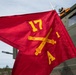 A red military guidon flag is displayed in the foreground, bearing gold crossed cannons (the U.S. Army Field Artillery branch insignia), the number '17', the number '1', and the letter 'A'. The flag indicates this is Alpha Battery, 1st Battalion, 17th Field Artillery Regiment. A military olive-drab vehicle is visible in the background.