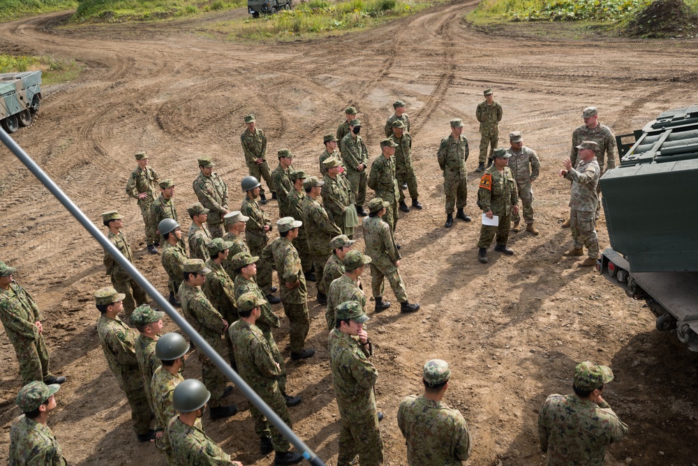 A large group of soldiers in camouflage uniforms are assembled on a dirt training ground, receiving a briefing or instruction near a military vehicle. Two instructors, appearing to wear a different camouflage pattern consistent with US Army uniforms, are addressing the assembled group. The setting appears to be a joint military training exercise, likely involving Japanese Ground Self-Defense Force personnel and US Army soldiers.