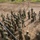 A large group of soldiers in camouflage uniforms are assembled on a dirt training ground, receiving a briefing or instruction near a military vehicle. Two instructors, appearing to wear a different camouflage pattern consistent with US Army uniforms, are addressing the assembled group. The setting appears to be a joint military training exercise, likely involving Japanese Ground Self-Defense Force personnel and US Army soldiers.