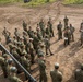 A large group of soldiers in camouflage uniforms are assembled on a dirt training ground, receiving a briefing or instruction near a military vehicle. Two instructors, appearing to wear a different camouflage pattern consistent with US Army uniforms, are addressing the assembled group. The setting appears to be a joint military training exercise, likely involving Japanese Ground Self-Defense Force personnel and US Army soldiers.