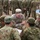 The image shows a joint military exercise or briefing between U.S. Army soldiers and what appear to be Japan Ground Self-Defense Force (JGSDF) soldiers. Two U.S. Army personnel in MultiCam/OCP uniforms are engaged in a ground-level discussion with several JGSDF soldiers wearing distinctive Japanese digital camouflage uniforms. A larger group of JGSDF soldiers stands in formation in the background.