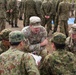 The image shows a joint military exercise or briefing between U.S. Army soldiers and what appear to be Japan Ground Self-Defense Force (JGSDF) soldiers. Two U.S. Army personnel in MultiCam/OCP uniforms are engaged in a ground-level discussion with several JGSDF soldiers wearing distinctive Japanese digital camouflage uniforms. A larger group of JGSDF soldiers stands in formation in the background.