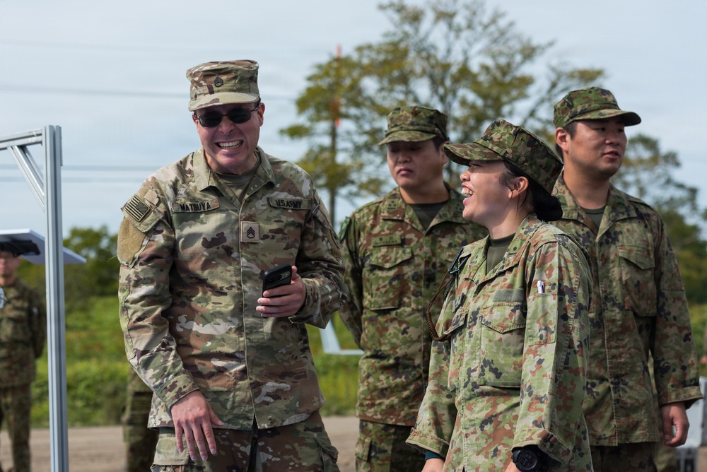 The image shows a group of military personnel from two different armed forces interacting in an outdoor setting. A U.S. Army soldier, identified by name tape 'MATBUYA' and U.S. Army insignia, is smiling and conversing with what appear to be Japan Ground Self-Defense Force (JGSDF) soldiers. The interaction appears friendly and relaxed, suggesting a joint training exercise or bilateral military engagement.