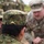 The image shows military personnel from what appear to be two different armed forces engaged in a briefing or discussion. The foreground soldier wears a Japanese Ground Self-Defense Force (JGSDF) camouflage uniform with a gold armor branch insignia on the cap, while the soldier in the background wears US Army Operational Camouflage Pattern (OCP) uniform. This appears to be a joint military exercise or training event.