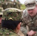 The image shows military personnel from what appear to be two different armed forces engaged in a briefing or discussion. The foreground soldier wears a Japanese Ground Self-Defense Force (JGSDF) camouflage uniform with a gold armor branch insignia on the cap, while the soldier in the background wears US Army Operational Camouflage Pattern (OCP) uniform. This appears to be a joint military exercise or training event.