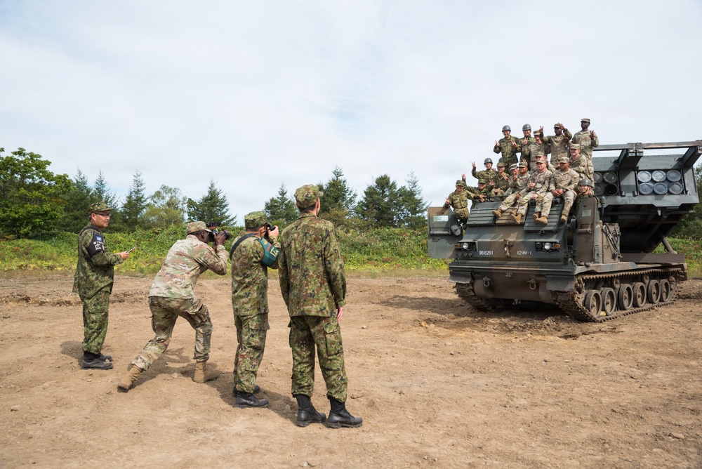 A group of military personnel from what appear to be two different nations are posing for a group photograph atop a tracked military vehicle, likely an M270 Multiple Launch Rocket System (MLRS) or similar platform. Photographers on the ground are capturing the moment while soldiers on the vehicle display peace signs and thumbs-up gestures. The setting appears to be a military training ground or exercise area.