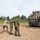 A group of military personnel from what appear to be two different nations are posing for a group photograph atop a tracked military vehicle, likely an M270 Multiple Launch Rocket System (MLRS) or similar platform. Photographers on the ground are capturing the moment while soldiers on the vehicle display peace signs and thumbs-up gestures. The setting appears to be a military training ground or exercise area.