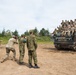A group of military personnel from what appear to be two different nations are posing for a group photograph atop a tracked military vehicle, likely an M270 Multiple Launch Rocket System (MLRS) or similar platform. Photographers on the ground are capturing the moment while soldiers on the vehicle display peace signs and thumbs-up gestures. The setting appears to be a military training ground or exercise area.