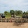 A large group of military personnel from what appears to be two different nations (indicated by two distinct camouflage uniform patterns) pose for a formal group photograph on a dirt field. They are positioned in front of multiple heavy artillery and rocket systems, including what appear to be MLRS/HIMARS-type rocket launchers and a self-propelled howitzer. A red unit guidon/pennant is visible being held in the center of the formation.