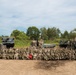 A large group of military personnel from what appears to be two different nations (indicated by two distinct camouflage uniform patterns) pose for a formal group photograph on a dirt field. They are positioned in front of multiple heavy artillery and rocket systems, including what appear to be MLRS/HIMARS-type rocket launchers and a self-propelled howitzer. A red unit guidon/pennant is visible being held in the center of the formation.