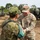 A U.S. Army soldier identified by name tape as 'QUINONES' interacts with a Japan Ground Self-Defense Force (JGSDF) soldier, appearing to exchange or present an item. Both are in their respective camouflage uniforms on what appears to be a military training or exercise area. In the background, military vehicles including what appears to be a self-propelled howitzer and a HIMARS or similar rocket artillery system are visible.