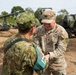A U.S. Army soldier identified by name tape as 'QUINONES' interacts with a Japan Ground Self-Defense Force (JGSDF) soldier, appearing to exchange or present an item. Both are in their respective camouflage uniforms on what appears to be a military training or exercise area. In the background, military vehicles including what appears to be a self-propelled howitzer and a HIMARS or similar rocket artillery system are visible.
