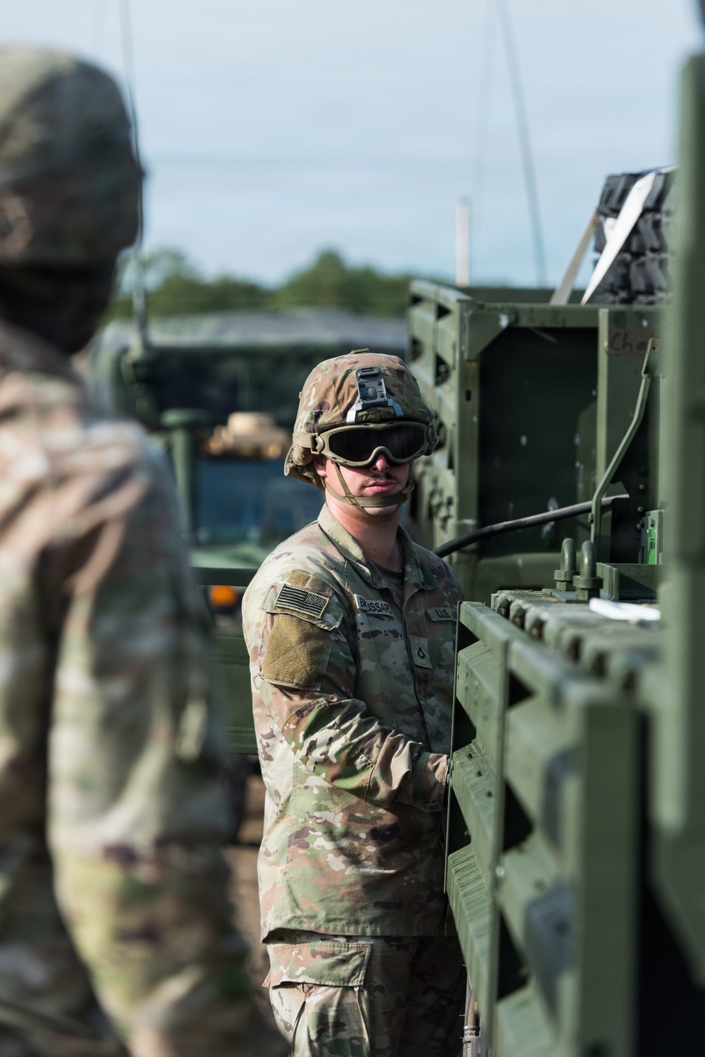 A U.S. Army soldier in Operational Camouflage Pattern (OCP) uniform is operating or inspecting military equipment, likely a tactical vehicle or weapons system. The soldier is wearing a combat helmet with goggles and has a U.S. flag patch visible on the right sleeve. A second soldier is partially visible in the foreground, out of focus.