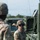 A U.S. Army soldier in Operational Camouflage Pattern (OCP) uniform is operating or inspecting military equipment, likely a tactical vehicle or weapons system. The soldier is wearing a combat helmet with goggles and has a U.S. flag patch visible on the right sleeve. A second soldier is partially visible in the foreground, out of focus.