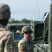 A U.S. Army soldier in Operational Camouflage Pattern (OCP) uniform is operating or inspecting military equipment, likely a tactical vehicle or weapons system. The soldier is wearing a combat helmet with goggles and has a U.S. flag patch visible on the right sleeve. A second soldier is partially visible in the foreground, out of focus.