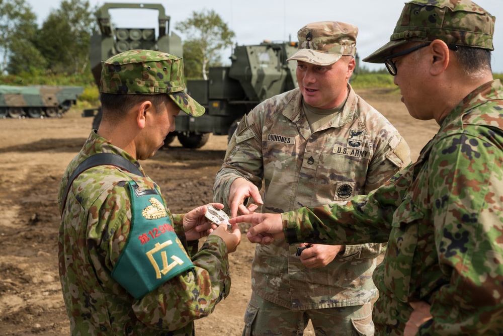 A U.S. Army soldier identified as 'QUINONES' interacts with two Japan Ground Self-Defense Force (JGSDF) soldiers in what appears to be a joint military exercise or exchange. The soldier is showing or handing a small object to the Japanese personnel. A military rocket artillery system, likely an M270 MLRS or HIMARS, is visible in the background.