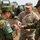 A U.S. Army soldier identified as 'QUINONES' interacts with two Japan Ground Self-Defense Force (JGSDF) soldiers in what appears to be a joint military exercise or exchange. The soldier is showing or handing a small object to the Japanese personnel. A military rocket artillery system, likely an M270 MLRS or HIMARS, is visible in the background.