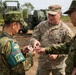 A U.S. Army soldier identified as 'QUINONES' interacts with two Japan Ground Self-Defense Force (JGSDF) soldiers in what appears to be a joint military exercise or exchange. The soldier is showing or handing a small object to the Japanese personnel. A military rocket artillery system, likely an M270 MLRS or HIMARS, is visible in the background.