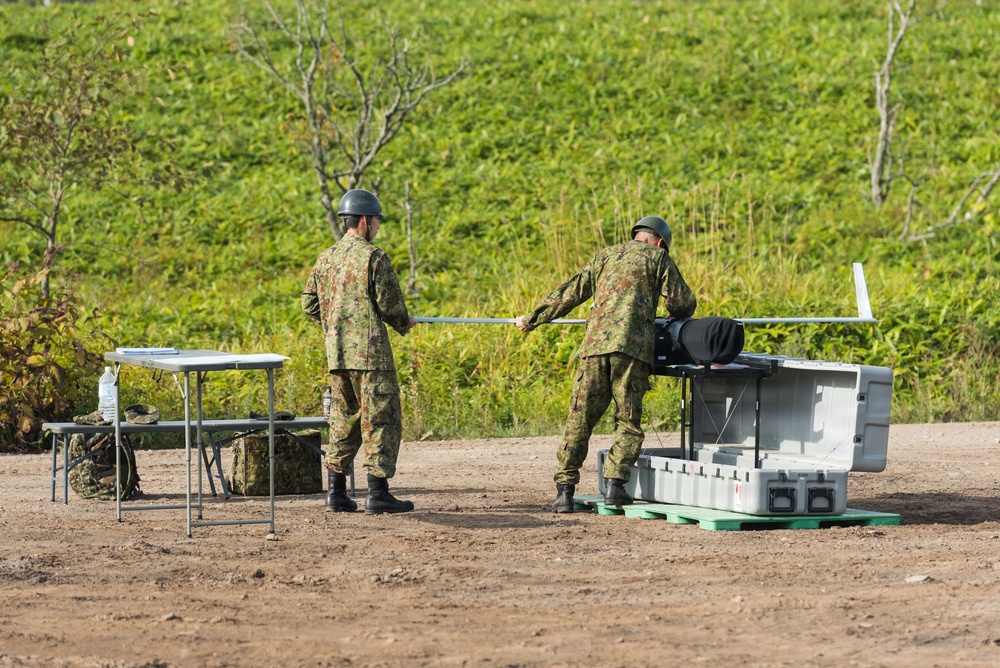 Two military personnel in camouflage uniforms and helmets are handling what appears to be a fixed-wing UAV (unmanned aerial vehicle) near its transport/launch case on a dirt clearing. The soldiers appear to be assembling or preparing the drone for operation, with a folding table set up nearby. The setting appears to be an outdoor military training or operational area with green vegetation in the background.