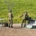 Two military personnel in camouflage uniforms and helmets are handling what appears to be a fixed-wing UAV (unmanned aerial vehicle) near its transport/launch case on a dirt clearing. The soldiers appear to be assembling or preparing the drone for operation, with a folding table set up nearby. The setting appears to be an outdoor military training or operational area with green vegetation in the background.