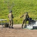 Two military personnel in camouflage uniforms and helmets are handling what appears to be a fixed-wing UAV (unmanned aerial vehicle) near its transport/launch case on a dirt clearing. The soldiers appear to be assembling or preparing the drone for operation, with a folding table set up nearby. The setting appears to be an outdoor military training or operational area with green vegetation in the background.