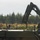 A military personnel is signaling with a raised hand near a military flatbed truck equipped with a hydraulic knuckle-boom crane. The scene appears to be an outdoor field operation, likely involving loading or unloading of equipment. The background features a treeline of conifers and low-lying vegetation consistent with a training or operational area.