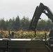 A military personnel is signaling with a raised hand near a military flatbed truck equipped with a hydraulic knuckle-boom crane. The scene appears to be an outdoor field operation, likely involving loading or unloading of equipment. The background features a treeline of conifers and low-lying vegetation consistent with a training or operational area.