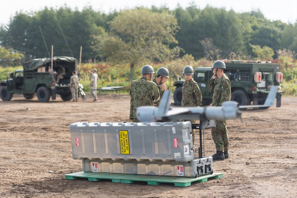 Military personnel in camouflage uniforms and helmets are gathered at an outdoor field site, appearing to conduct operations or training exercises. In the foreground, a large gray equipment case with a 'DO NOT SIT ON THIS SURFACE' warning label sits on a green pallet, with what appears to be a small UAV or drone mounted on a stand nearby. Multiple military vehicles are visible in the background along with additional personnel.