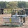 Military personnel in camouflage uniforms and helmets are gathered at an outdoor field site, appearing to conduct operations or training exercises. In the foreground, a large gray equipment case with a 'DO NOT SIT ON THIS SURFACE' warning label sits on a green pallet, with what appears to be a small UAV or drone mounted on a stand nearby. Multiple military vehicles are visible in the background along with additional personnel.