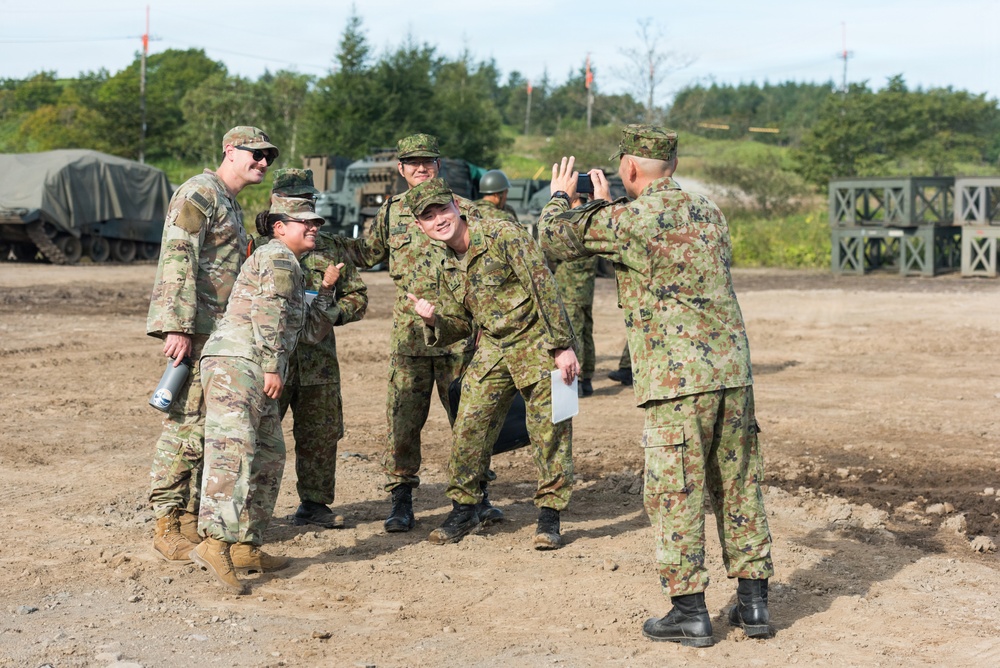 The image shows a group of military personnel from two different armed forces posing together for a photograph at what appears to be a joint military exercise or training area. Soldiers wearing U.S. Army MultiCam (OCP) uniforms are interacting with soldiers wearing Japanese Ground Self-Defense Force (JGSDF) woodland camouflage uniforms. The atmosphere appears informal and friendly, suggesting a cooperative bilateral military engagement.