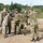The image shows a group of military personnel from two different armed forces posing together for a photograph at what appears to be a joint military exercise or training area. Soldiers wearing U.S. Army MultiCam (OCP) uniforms are interacting with soldiers wearing Japanese Ground Self-Defense Force (JGSDF) woodland camouflage uniforms. The atmosphere appears informal and friendly, suggesting a cooperative bilateral military engagement.