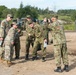 The image shows a group of military personnel from two different armed forces posing together for a photograph at what appears to be a joint military exercise or training area. Soldiers wearing U.S. Army MultiCam (OCP) uniforms are interacting with soldiers wearing Japanese Ground Self-Defense Force (JGSDF) woodland camouflage uniforms. The atmosphere appears informal and friendly, suggesting a cooperative bilateral military engagement.