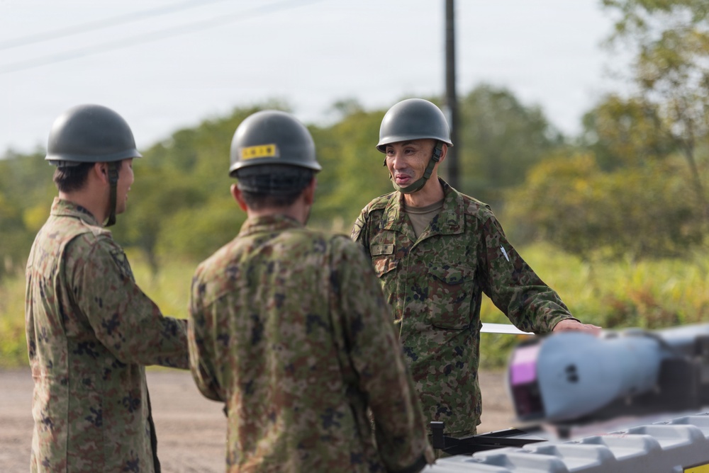 Three military personnel in camouflage uniforms and gray helmets are engaged in a discussion in an outdoor field setting. The soldier facing the camera appears to be briefing or instructing the other two. Military equipment, possibly munitions or ordnance, is visible in the foreground.