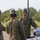 Three military personnel in camouflage uniforms and gray helmets are engaged in a discussion in an outdoor field setting. The soldier facing the camera appears to be briefing or instructing the other two. Military equipment, possibly munitions or ordnance, is visible in the foreground.