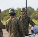 Three military personnel in camouflage uniforms and gray helmets are engaged in a discussion in an outdoor field setting. The soldier facing the camera appears to be briefing or instructing the other two. Military equipment, possibly munitions or ordnance, is visible in the foreground.