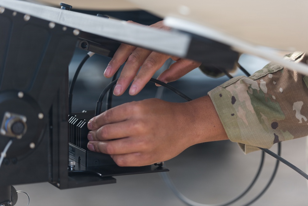 A military service member in MultiCam/OCP camouflage uniform is working on electronic equipment mounted inside what appears to be an aircraft or military vehicle panel. The person is using both hands to handle or install a black electronic component with heat sink fins and cable connections. The close-up shot focuses on the hands and the equipment being serviced.