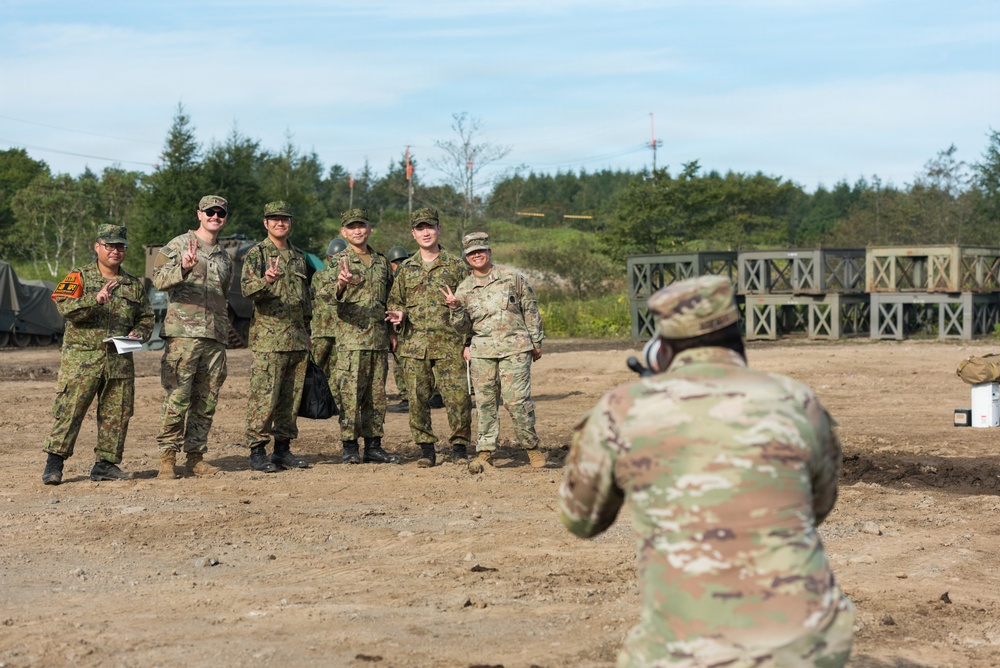 A group of military personnel from at least two different nations pose for a group photograph at what appears to be a joint military training exercise or field operation. Several soldiers are making peace/victory hand signs while smiling, suggesting a friendly, cooperative atmosphere. A soldier in the foreground is photographing the group using a camera.