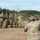 A group of military personnel from at least two different nations pose for a group photograph at what appears to be a joint military training exercise or field operation. Several soldiers are making peace/victory hand signs while smiling, suggesting a friendly, cooperative atmosphere. A soldier in the foreground is photographing the group using a camera.