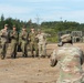 A group of military personnel from at least two different nations pose for a group photograph at what appears to be a joint military training exercise or field operation. Several soldiers are making peace/victory hand signs while smiling, suggesting a friendly, cooperative atmosphere. A soldier in the foreground is photographing the group using a camera.
