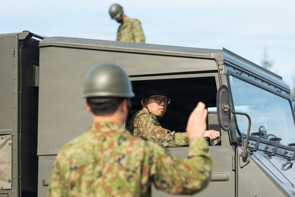 The image shows military personnel in camouflage uniforms and olive-drab helmets operating a military vehicle. One soldier is seated in the driver's position of a green tactical vehicle, looking out the open window, while another soldier in the foreground appears to be directing or signaling with a raised hand. A third soldier is visible standing on or behind the vehicle in the background.