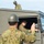 The image shows military personnel in camouflage uniforms and olive-drab helmets operating a military vehicle. One soldier is seated in the driver's position of a green tactical vehicle, looking out the open window, while another soldier in the foreground appears to be directing or signaling with a raised hand. A third soldier is visible standing on or behind the vehicle in the background.