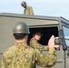 The image shows military personnel in camouflage uniforms and olive-drab helmets operating a military vehicle. One soldier is seated in the driver's position of a green tactical vehicle, looking out the open window, while another soldier in the foreground appears to be directing or signaling with a raised hand. A third soldier is visible standing on or behind the vehicle in the background.