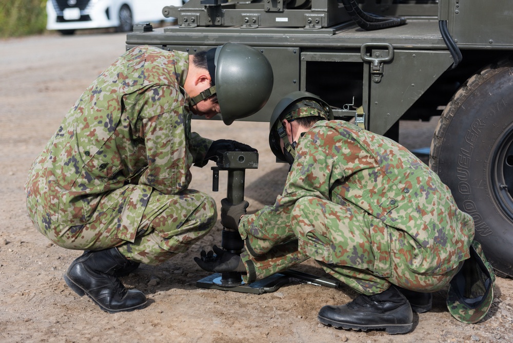 Two military personnel in camouflage uniforms and helmets are crouching beside a military vehicle, working on what appears to be a stabilizer jack or outrigger mechanism on the ground. The soldiers are wearing Japanese Ground Self-Defense Force (JGSDF) distinctive woodland camouflage pattern uniforms. The activity appears to be equipment setup or maintenance in an outdoor training or operational environment.
