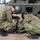 Two military personnel in camouflage uniforms and helmets are crouching beside a military vehicle, working on what appears to be a stabilizer jack or outrigger mechanism on the ground. The soldiers are wearing Japanese Ground Self-Defense Force (JGSDF) distinctive woodland camouflage pattern uniforms. The activity appears to be equipment setup or maintenance in an outdoor training or operational environment.
