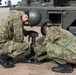 Two military personnel in camouflage uniforms and helmets are crouching beside a military vehicle, working on what appears to be a stabilizer jack or outrigger mechanism on the ground. The soldiers are wearing Japanese Ground Self-Defense Force (JGSDF) distinctive woodland camouflage pattern uniforms. The activity appears to be equipment setup or maintenance in an outdoor training or operational environment.