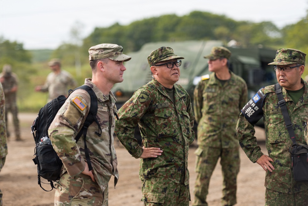 The image shows a joint military gathering involving U.S. Army personnel and what appear to be Japan Ground Self-Defense Force (JGSDF) soldiers, likely during a bilateral training exercise or field operation. The U.S. soldier on the left wears OCP (Operational Camouflage Pattern) with a visible American flag patch, while the other personnel wear a distinctive woodland camouflage pattern consistent with JGSDF uniforms. The setting appears to be an outdoor training area with military vehicles visible in the background.
