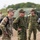 The image shows a joint military gathering involving U.S. Army personnel and what appear to be Japan Ground Self-Defense Force (JGSDF) soldiers, likely during a bilateral training exercise or field operation. The U.S. soldier on the left wears OCP (Operational Camouflage Pattern) with a visible American flag patch, while the other personnel wear a distinctive woodland camouflage pattern consistent with JGSDF uniforms. The setting appears to be an outdoor training area with military vehicles visible in the background.