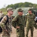 The image shows a joint military gathering involving U.S. Army personnel and what appear to be Japan Ground Self-Defense Force (JGSDF) soldiers, likely during a bilateral training exercise or field operation. The U.S. soldier on the left wears OCP (Operational Camouflage Pattern) with a visible American flag patch, while the other personnel wear a distinctive woodland camouflage pattern consistent with JGSDF uniforms. The setting appears to be an outdoor training area with military vehicles visible in the background.